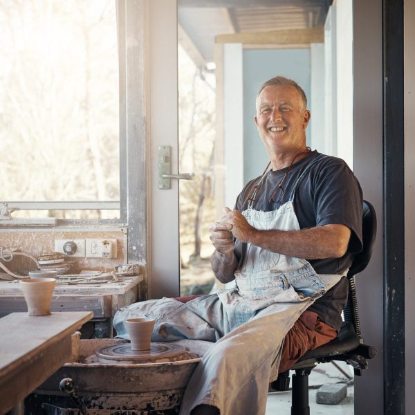 A white man wearing a dark grey shirt and white apron with short blond hair sits making pottery.