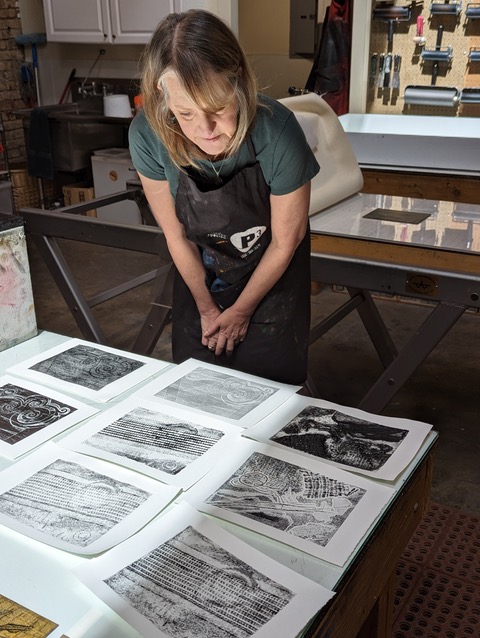 A woman in an artist's apron looks at an array of black-and-white prints.